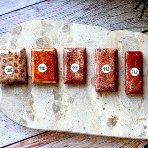 May include: Five rectangular coral pendants in shades of red and orange, each with a white sticker displaying a number from 109 to 113. The pendants are displayed on a light grey, speckled surface.
