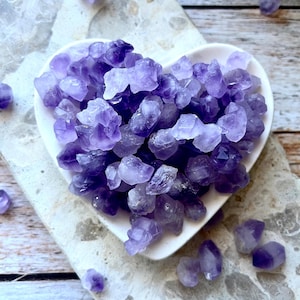 May include: A collection of raw, deep purple amethyst crystal shards contained within a white, heart-shaped ceramic dish. The stones vary in size and transparency, displaying natural jagged edges and multifaceted surfaces in shades of violet.