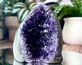 May include: A natural amethyst geode featuring a deep purple, crystalline interior with sharp, pointed points. The exterior of the stone is a rough, grey rock formation, showcasing a hollow center filled with sparkling, translucent violet crystals.
