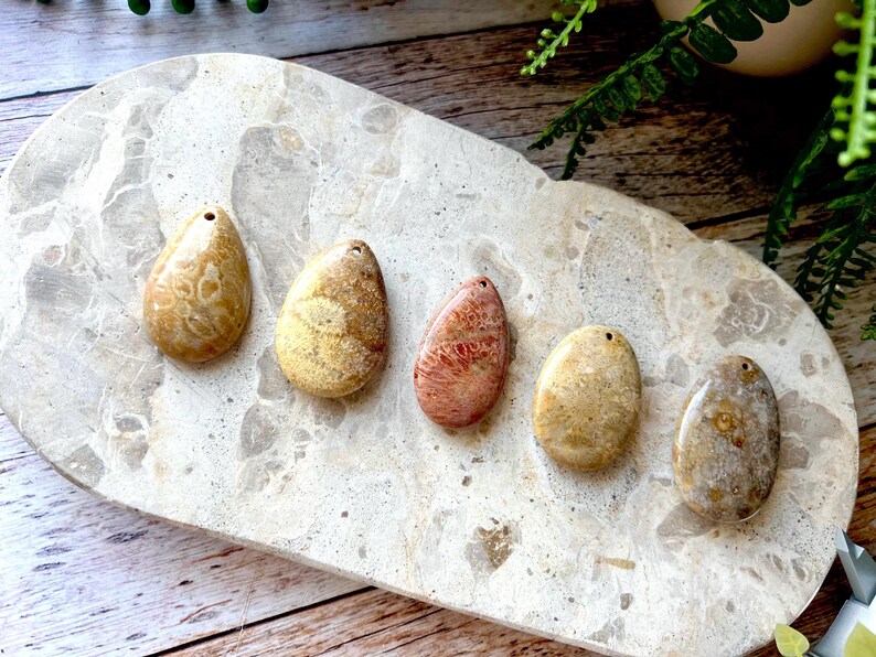 May include: Five polished, teardrop-shaped stones in shades of beige, tan, and coral are displayed on a light-coloured, textured surface. Each stone has a small hole drilled at the top. The stones vary in pattern and colour.
