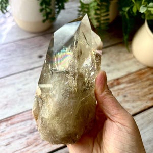 May include: A large, translucent smoky quartz crystal point with a natural, rough base and a polished, faceted top. The crystal displays internal fractures and a distinct rainbow-colored light refraction near the apex of the stone.