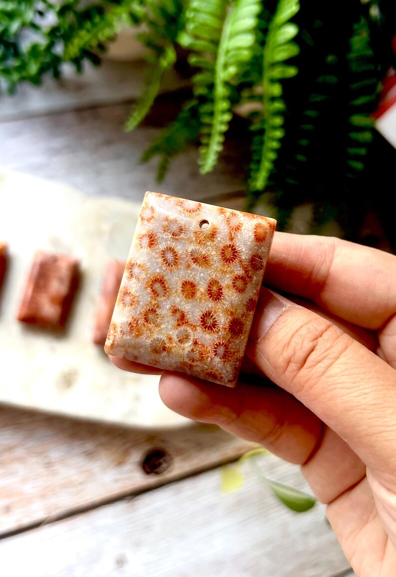 May include: A rectangular coral pendant with a hole at the top. The pendant features a pattern of small, orange-red flower-like shapes on a cream-coloured background. The pendant is held in a hand, with other coral pieces visible in the background.