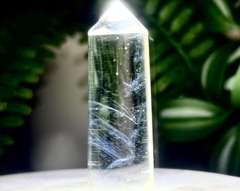 May include: A clear, cylindrical quartz crystal point with a faceted top. The transparent stone features internal white and blue wispy inclusions that resemble delicate, feathery patterns suspended within the solid, polished mineral structure.