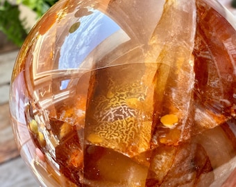 May include: A polished, spherical quartz crystal featuring internal inclusions of deep amber and burnt orange mineral deposits. The smooth, translucent surface displays complex, layered patterns and natural fractures within the stone's core.