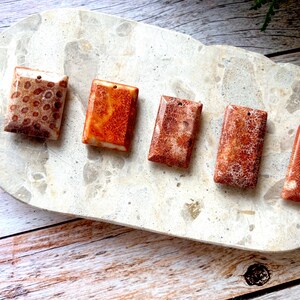 May include: Five rectangular coral pendants in shades of brown and orange are displayed on a light-coloured, oval-shaped stone tray. Each pendant has a small hole at the top. The pendants have unique patterns and textures.