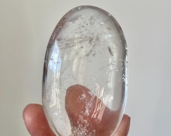 May include: A clear, oval-shaped crystal with internal inclusions. The polished stone is held in a hand, showcasing its transparency and natural beauty. The crystal's surface reflects light, highlighting its smooth texture. A decorative item.