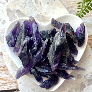 May include: A collection of raw amethyst crystals in varying shades of purple and black, displayed in a white heart-shaped dish. The crystals have sharp, jagged edges and a natural, unpolished appearance. The dish sits on a light-colored surface.