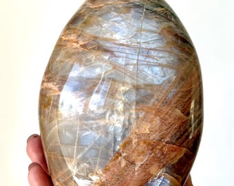 May include: A polished, oval-shaped gemstone with a mix of brown, tan, and gray hues. The stone has a smooth, glossy surface with visible patterns and lines. The gemstone is held in a hand, showcasing its size and shape.