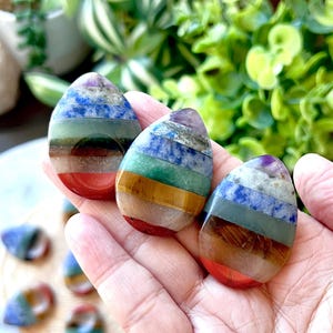 Puede incluir: Tres piedras de chakra en forma de lágrima en una mano. Cada piedra presenta capas de rojo, naranja, amarillo, verde, azul y morado. Se ven piedras adicionales en el fondo, junto con follaje verde.