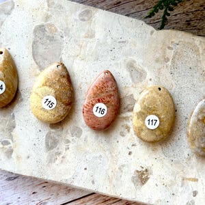 May include: Five teardrop-shaped pendants in shades of tan, beige, and coral, each with a small hole at the top. Each pendant has a white sticker with a number. The pendants are displayed on a light-coloured, textured surface.