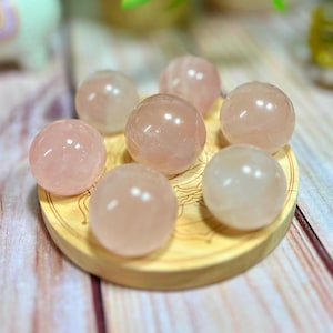 May include: Seven smooth, pink rose quartz spheres arranged on a wooden tray. The spheres are polished and translucent, with a slight pink hue.