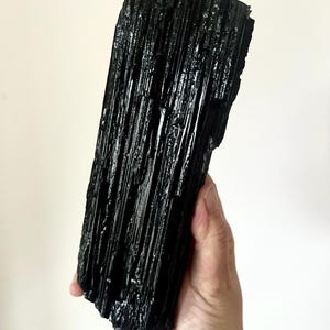May include: A large, elongated black tourmaline crystal specimen featuring a deeply striated, vertical texture with a glossy, reflective surface. The mineral has a raw, jagged top edge and a solid, opaque appearance with natural crystalline formations.