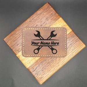 May include: A brown leatherette patch with the text "Your Name Here" and a crossed wrench design. The patch is stitched onto a wooden coaster with a diamond shape and a dark background.