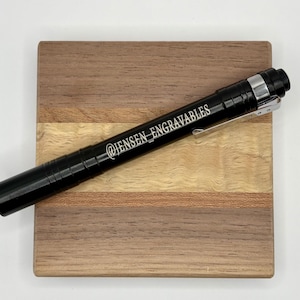May include: A black metal pen with a silver clip and the text "@JENSEN_ENGRAVABLES" engraved on the side. The pen is lying on a wooden surface with a light brown and dark brown striped pattern.
