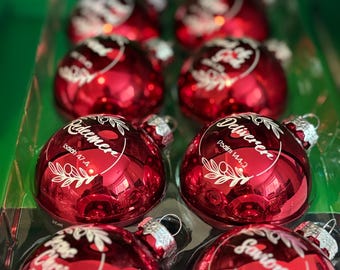Puede incluir: Un juego de adornos navideños rojos con tapas plateadas. Cada adorno tiene una escritura blanca y un diseño floral. El texto en los adornos incluye las palabras "Redeemer", "Deliverer", "Prince Christ" y "Savior".