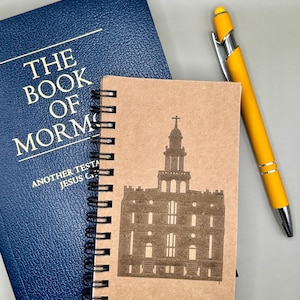 Puede incluir: Un cuaderno marrón de espiral con una silueta del Templo de Salt Lake en la portada. El cuaderno está al lado de un libro azul con el texto "El Libro de Mormón" en la portada. Un bolígrafo amarillo está sobre la mesa.
