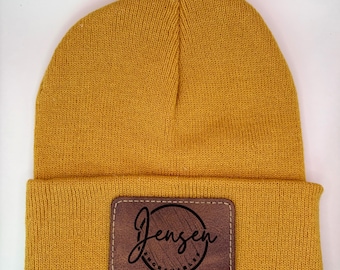 May include: A mustard yellow beanie with a brown leather patch that says "Jensen" in a circle with a wood grain texture.