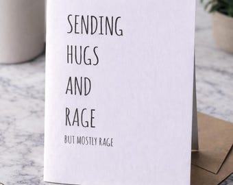 Puede incluir: Una tarjeta de felicitación blanca con el texto "SENDING HUGS AND RAGE BUT MOSTLY RAGE" en negro. La tarjeta está sobre una superficie de mármol, con una taza de café, una planta y un bloc de notas con un lápiz en el fondo.