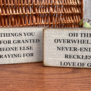 Op de afbeelding: Twee rechthoekige houten borden met tekst op een achtergrond in krantenstijl. Op één bord staat "THE THINGS YOU TAKE FOR GRANTED SOMEONE ELSE IS PRAYING FOR." Op de andere staat "OH THE OVERWHELMING NEVER-ENDING RECKLESS LOVE OF GOD."