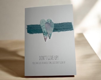 Pode incluir: Um cartão cinza com dois corações azuis e roxos sobre uma fita turquesa. O texto "DON'T GIVE UP!" e uma citação de apoio são impressos abaixo. Um cartão motivacional.