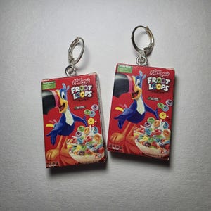 May include: A pair of earrings featuring miniature Kellogg's Froot Loops cereal boxes. The boxes are red with the Froot Loops logo and a toucan mascot. The earrings have silver-colored leverback closures.