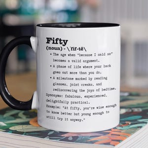 May include: White ceramic mug with a black handle and rim, featuring the definition of "Fifty." The text includes synonyms and an example sentence. The mug is on a book with a floral design.