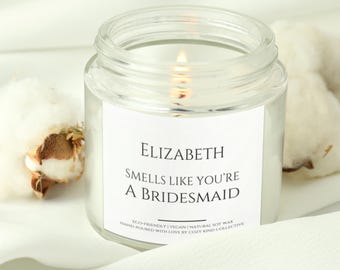 Może przedstawiać: Szklany słoik z przezroczystego szkła z białą etykietą z napisem "ELIZABETH SMELLS LIKE YOU'RE A BRIDESMAID ECO-FRIENDLY | VEGAN | NATURAL SOY WAX HAND POURED WITH LOVE BY COZY KIND COLLECTIVE". Świeca jest zapalona i płomień jest widoczny.