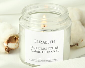 Könnte beinhalten: Eine Kerze in einem durchsichtigen Glasgefäß mit einem weißen Etikett, auf dem "ELIZABETH SMELLS LIKE YOU'RE A MAID OF HONOR" und "ECO-FRIENDLY | VEGAN | NATURAL SOY WAX HAND POURED WITH LOVE BY COZY KIND COLLECTIVE" steht. Die Kerze ist angezündet und die Flamme ist sichtbar.