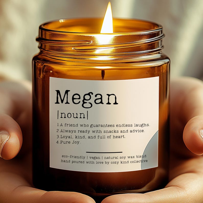 May include: A brown glass candle jar with a white label that reads "Megan noun 1. A friend who guarantees endless laughs. 2. Always ready with snacks and advice. 3. Loyal, kind, and full of heart. 4. Pure Joy eco-friendly | vegan | natural soy wax blend hand poured with love by cozy kind collective". The candle is lit and the flame is visible.