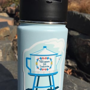 May include: A light blue water bottle with a sticker of a blue water tower with a white roof and a floral wreath around the words "Välkommen till Lindstrom".