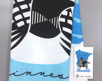 May include: White tea towel with a blue and black loon design. The towel has the word "Minnesota" written in black script. A small tag is attached to the towel with a loon and the words "Minnesota Tea Towel".