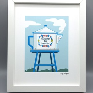 May include: A white framed print of a blue and white water tower shaped like a teapot with the text "Välkommen till Lindström" in a floral wreath. The print has a blue sky background with white clouds and green grass.
