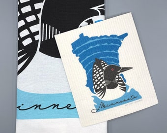 May include: A white kitchen towel with a blue and black loon design. The towel has the word "Minnesota" written on it. The towel is folded in half and is sitting on a gray surface.