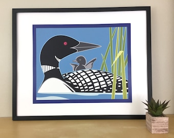 May include: Framed art print featuring a stylized illustration of a loon with a chick. The loon is black, white, and gray, with a red eye, and the chick is gray. The background is blue with green reeds.