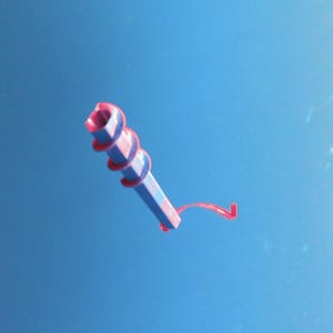 May include: A blue and pink decorative item with a spiral design and a red string. The item has a hexagonal shape and a hollow center. The background is a solid blue.