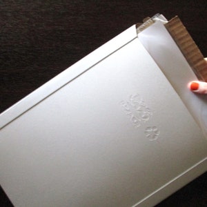 Op de afbeelding: Een witte kartonnen envelop met een reli&euml;fdesign van een gestileerde bloem. De envelop is gevouwen en heeft een flap aan de bovenkant.