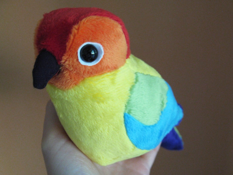 K&ouml;nnte beinhalten: Ein Pl&uuml;schtier in Form eines Vogels mit roten, orangen, gelben, gr&uuml;nen, blauen und violetten Federn. Der Vogel hat einen schwarzen Schnabel und gro&szlig;e schwarze Augen.