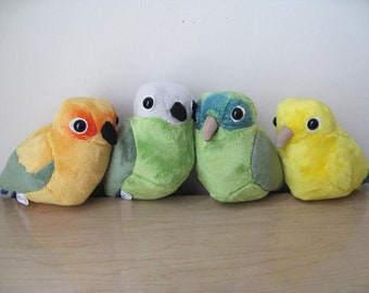 May include: Four plush toy birds in different colors. The birds are green, yellow, gray, and orange. They are all sitting on a wooden surface.