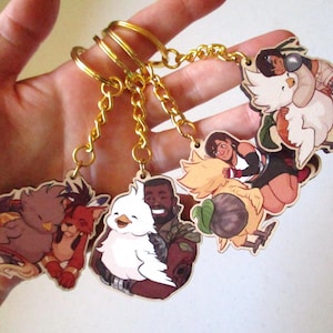 May include: Five keychains featuring cartoon characters with a white bird. Each keychain has a gold chain and a different character. The characters include a red-haired woman, a man with brown hair, a woman with black hair, a man with brown skin, and a white bird.