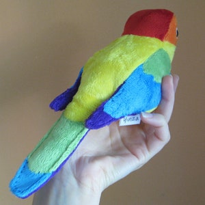 K&ouml;nnte beinhalten: Ein Pl&uuml;schtier in Form eines Regenbogen-Vogels mit rotem Kopf und einem lilafarbenen Etikett mit der Aufschrift "Yinza".