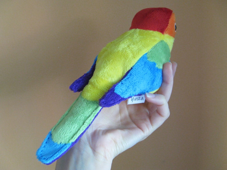 K&ouml;nnte beinhalten: Ein Pl&uuml;schtier in Form eines Regenbogen-Vogels mit rotem Kopf und einem lilafarbenen Etikett mit der Aufschrift "Yinza".