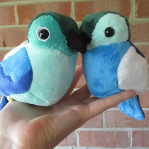 May include: Two plush bird toys in shades of blue, teal, and white are held in a hand. The birds have black beaks and large black eyes. The birds are made of soft fabric and are likely handmade.