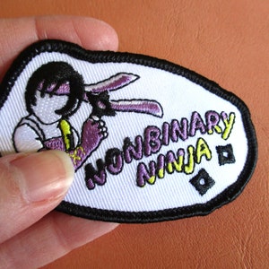 Puede incluir: Parche blanco bordado con un dibujo animado de una persona con ropa morada y amarilla y el texto "Nonbinary Ninja" en letras moradas y amarillas.