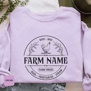 May include: A lavender sweatshirt with a black and white farm logo. The logo features a chicken, leaves, and the text "ESTD - 2025", "FARM NAME", "FARM FRESH", "EGGS", "VEGETABLES", and "STRAW".