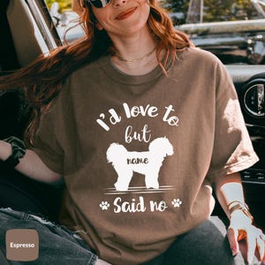 May include: A brown t-shirt with the text "I'd love to but [name] said no" and a white silhouette of a dog. The text is in a white font. The dog is facing right. The t-shirt is on a person.