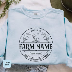 May include: Light blue sweatshirt with a black and white farm logo. The logo features a chicken, the text "ESTD - 2025", "FARM NAME", "FARM FRESH", and "EGGS - VEGETABLES - STRAW".