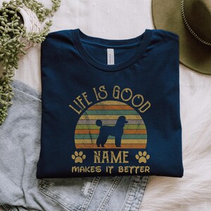May include: A navy blue t-shirt with a vintage sunset graphic and the text "Life is good NAME makes it better". A silhouette of a poodle is in front of the sunset.  Two paw prints are on either side of the text "NAME".