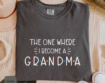 May include: Dark gray t-shirt with the text "THE ONE WHERE I BECOME A GRANDMA" in white. The word "GRANDMA" has colorful dots. The shirt is made of a soft material and is a Comfort Colors brand.