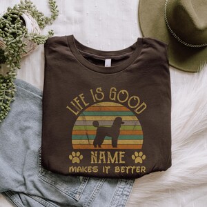 May include: A brown t-shirt with a vintage-style sunset graphic and a silhouette of a poodle. The text "Life is good" is above the sunset and "NAME makes it better" is below.