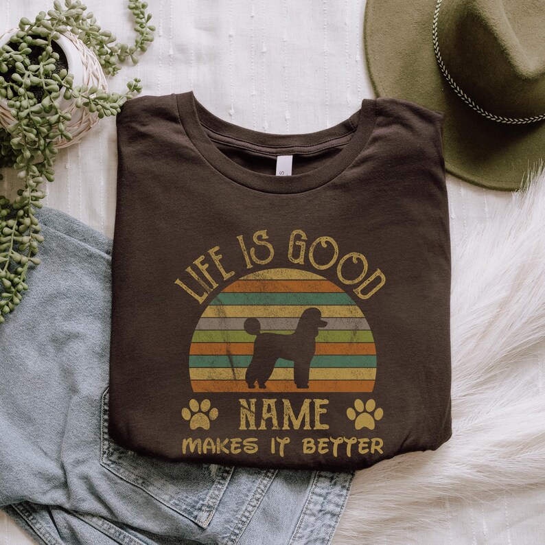 May include: A brown t-shirt with a vintage-style sunset graphic and a silhouette of a poodle. The text "Life is good" is above the sunset and "NAME makes it better" is below.